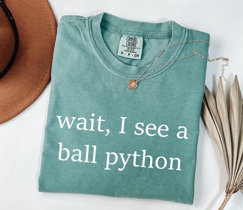 K&ouml;nnte beinhalten: Ein kurz&auml;rmeliges T-Shirt in Salbeigr&uuml;n mit Rundhalsausschnitt und dem wei&szlig;en Text "wait, I see a ball python" auf der Vorderseite. Das Shirt hat ein Comfort Colors Markenetikett am Innenkragen und besteht aus 100% Baumwollstoff.