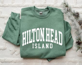 May include: A green crewneck sweatshirt with the words "HILTON HEAD ISLAND" in white block letters. The sweatshirt is folded neatly on a white textured surface. A sprig of faux pine and pinecones is in the background.