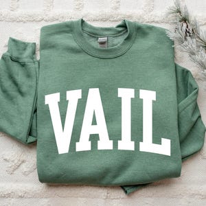 May include: A sage green crewneck sweatshirt with the word "VAIL" in large, white, block letters across the front. The sweatshirt is folded neatly on a white, textured surface. A sprig of faux evergreen and pinecones is in the background.