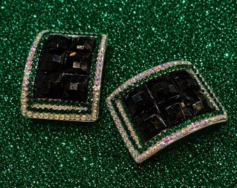 May include: Two square-shaped, silver-toned accessories with a black, geometric center. The edges are adorned with a row of green and iridescent rhinestones. The background is a sparkly, emerald green.