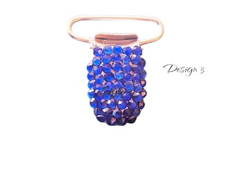 May include: A silver-toned metal clip with a cluster of blue faceted beads. The beads are arranged in a rectangular shape, and the clip has a curved handle. The text "Design 3" is written in cursive.