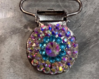 May include: A circular metal pacifier clip featuring a central iridescent crystal surrounded by a ring of light blue rhinestones and an outer layer of clear, multi-colored reflective crystals. The text Design A Light blue is printed below.
