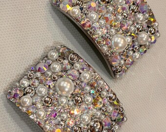 May include: Two rectangular hair clips densely embellished with a variety of white faux pearls, iridescent faceted crystals, and small silver metallic beads. The surface of each clip is completely covered in these mixed decorative elements.