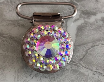 May include: A silver-toned pacifier clip with a circular, iridescent rhinestone design. The center features a large, multi-colored rhinestone surrounded by smaller, matching stones. The text "DESIGN 1" is printed below the clip.