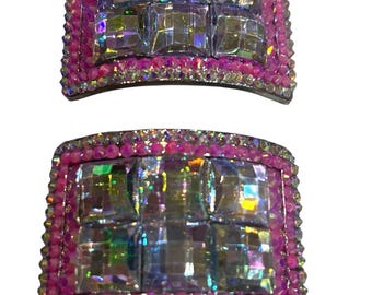 May include: Two decorative hair clips. Each clip features a rectangular design with a central arrangement of large, iridescent, square-cut crystals. The crystals are surrounded by a border of smaller, pink, sparkling rhinestones, and a pink frame.