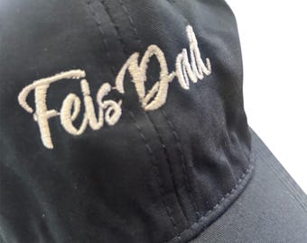 May include: A navy blue baseball cap with the embroidered text "Feis Dad" in a cursive font. The cap has a curved brim and a six-panel design, with visible stitching details.