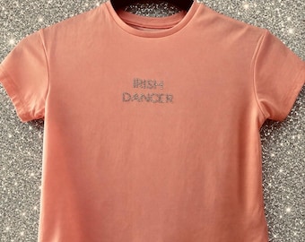 May include: A peach-colored t-shirt with the text "IRISH DANCER" in sparkling rhinestones. The short-sleeved shirt is hanging on a black hanger against a glittery background. A tag hangs from the bottom of the shirt.