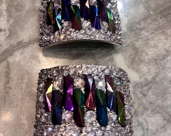 May include: Two silver-toned, rectangular hair clips. Each clip is encrusted with small, clear crystals around the edges. The center of each clip features a row of iridescent, multi-colored, geometric-shaped crystals, reflecting hues of blue, green, purple, and gold.