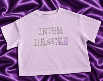 May include: A light purple short-sleeved t-shirt featuring the text IRISH DANCER spelled out in small, reflective silver-toned rhinestones across the chest. The garment has a crew neckline and a straight hemline, showcasing a simple design.