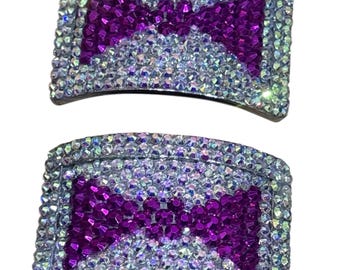 May include: Two silver-toned hair clips encrusted with clear and iridescent rhinestones. Each clip features a bow design in vibrant purple rhinestones, creating a sparkling and glamorous accessory.