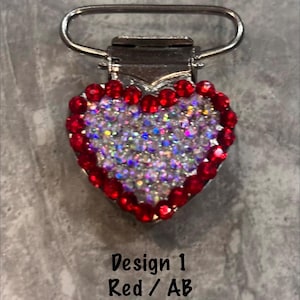 May include: A heart-shaped dummy clip with a silver metal clasp and loop. The heart is outlined with red rhinestones and filled with iridescent AB rhinestones. The text "Design 1 Red / AB" is below the heart.