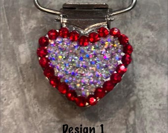 May include: A heart-shaped pacifier clip with a silver metal clasp and loop. The heart is outlined with red rhinestones and filled with iridescent AB rhinestones. The text "Design 1 Red / AB" is below the heart.