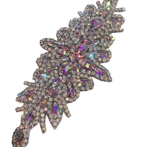 May include: An elaborate, floral-shaped applique. The design is covered in iridescent rhinestones in silver, purple, and clear. The central flower shape is surrounded by leaves. This decorative embellishment is suitable for adding sparkle to clothing or accessories.