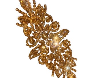 May include: An elongated decorative applique featuring a dense arrangement of amber-colored crystals and rhinestones in various shapes, including marquise and round cuts, set in a gold-toned metal frame with a symmetrical, ornate floral pattern.