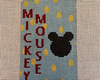 May include: A blue and white canvas needlepoint ornament with yellow polka dots. The ornament is shaped like a tag and has the text "Mickey Mouse" written vertically in red letters.
