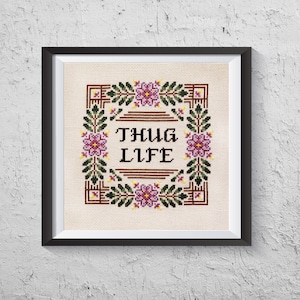 May include: Framed cross-stitch art with the words "THUG LIFE" in black, centered within a floral wreath. The wreath features purple flowers, green leaves, and brown geometric accents. The artwork is framed in a black square frame.