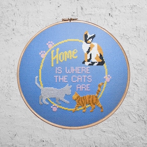 Op de afbeelding: Een blauwe borduurring met een kruissteekontwerp met drie katten en de tekst "Home is where the cats are".