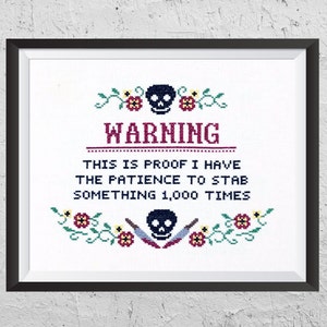 Op de afbeelding: Een zwart-wit kruissteekontwerp met een schedel en gekruiste beenderen en de tekst "WARNING THIS IS PROOF I HAVE THE PATIENCE TO STAB SOMETHING 1,000 TIMES". Het ontwerp is ingelijst in een zwart frame.