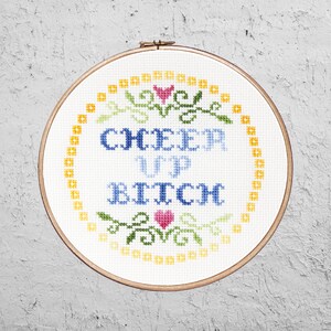 May include: A white embroidery hoop with a yellow border and a cross-stitch design that says "Cheer Up Bitch" with a green and pink floral design around the text.