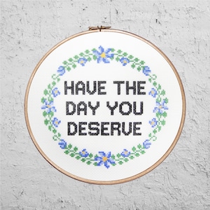 Puede incluir: Un aro de bordado blanco con un borde floral y el texto "HAVE THE DAY YOU DESERVE" en negro.