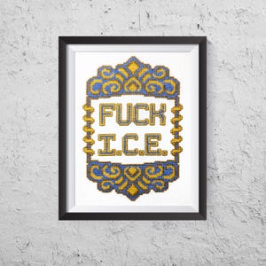 May include: Framed cross-stitch art with the words "FUCK ICE" in bold letters. The text is centered within a decorative blue and gold border. The artwork is displayed in a black frame against a textured white wall.
