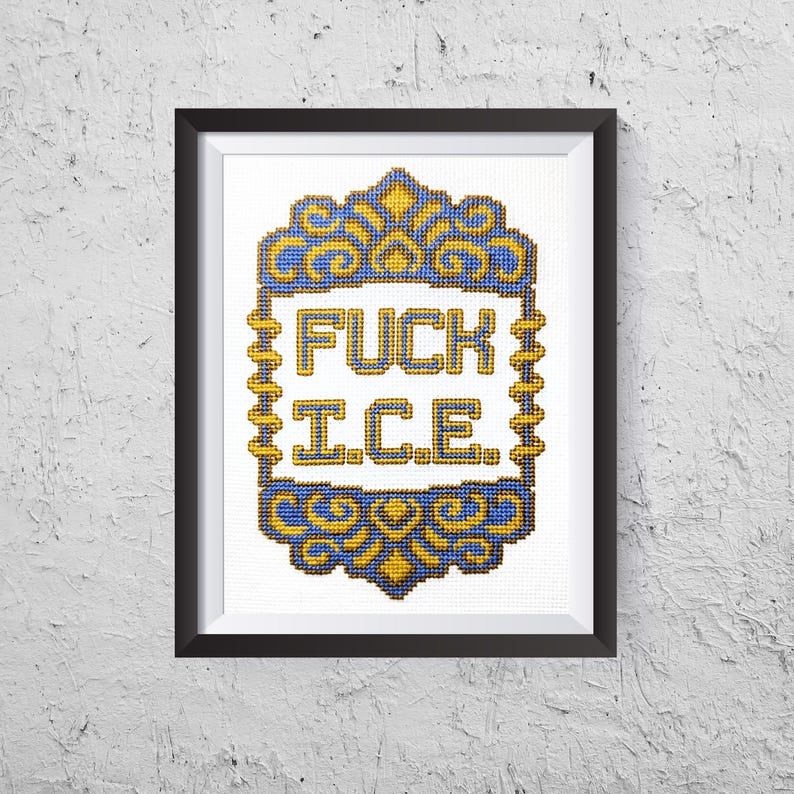 May include: Framed cross-stitch art with the words "FUCK ICE" in bold letters. The text is centered within a decorative blue and gold border. The artwork is displayed in a black frame against a textured white wall.