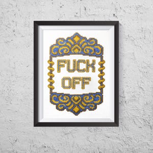 May include: Framed cross-stitch art with the words "FUCK OFF" in bold letters. The text is centered within a decorative blue and gold border. The artwork is framed in a black frame and mounted on a white textured wall.