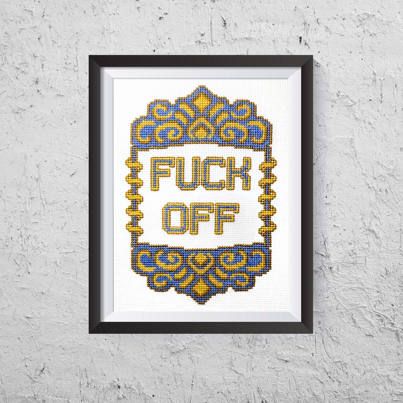 May include: Framed cross-stitch art with the words "FUCK OFF" in bold letters. The text is centered within a decorative blue and gold border. The artwork is framed in a black frame and mounted on a white textured wall.
