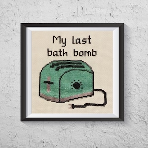 May include: A framed cross-stitch design featuring a green toaster with the text "My last bath bomb".