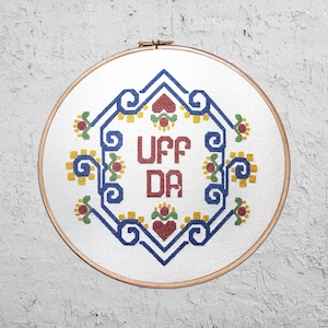 May include: A white embroidery hoop with a blue, red, and yellow geometric design. The words "UFF DA" are embroidered in red letters in the center of the design.