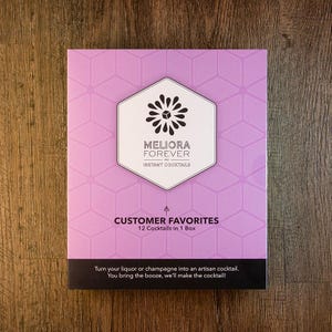 May include: A rectangular box with a light purple geometric pattern. The box features the text "MELIORA FOREVER INSTANT COCKTAILS" and "CUSTOMER FAVORITES 12 Cocktails in 1 Box". The bottom of the box reads "Turn your liquor or champagne into an artisan cocktail."