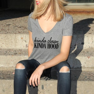 May include: A woman wearing a gray V-neck t-shirt with the text "kinda classy KINDA HOOD" printed on it. She is sitting on a set of concrete steps wearing blue jeans with rips in the knees.