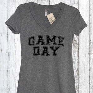 May include: A heather grey V-neck t-shirt with the text "GAME DAY" printed in black with a distressed look. The shirt has a tag that says "TYD Clothing".