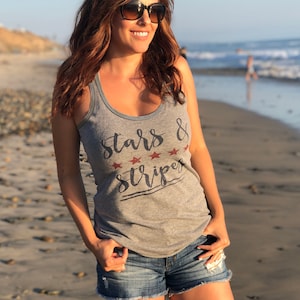 Op de afbeelding: Een grijze tanktop met de tekst "Stars & Stripes" in een scriptlettertype en rode sterren. De persoon draagt een spijkerbroek en een zonnebril op het strand. De foto is overdag gemaakt.