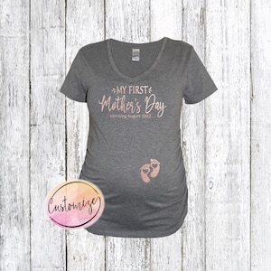 May include: Gray maternity shirt with a pink glitter design that says "My First Mother's Day - Arriving August 2022". There are two pink glitter baby footprints below the text.