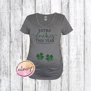 May include: A gray maternity t-shirt with the text "Extra lucky this year" and "Arriving August 2022". Two green four-leaf clovers are below the text.