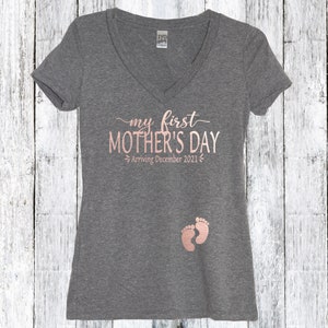 Puede incluir: Camiseta gris de cuello en V con el texto "my first MOTHER'S DAY Arriving December 2021" en letras doradas rosas. Dos huellas de pies de beb&eacute; rosas est&aacute;n impresas en la parte inferior de la camiseta.