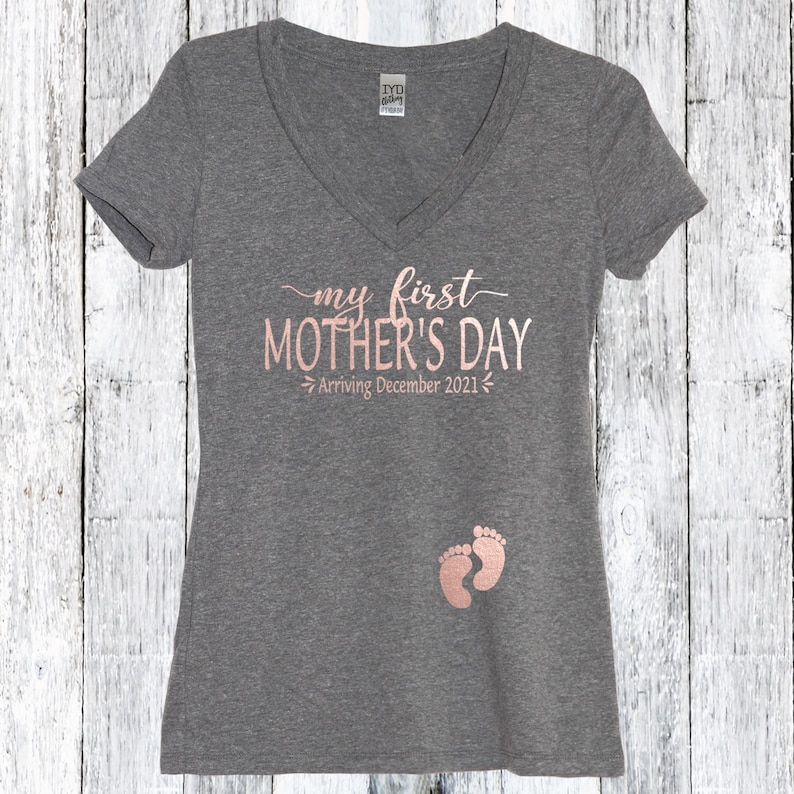 Puede incluir: Camiseta gris de cuello en V con el texto "my first MOTHER'S DAY Arriving December 2021" en letras doradas rosas. Dos huellas de pies de beb&eacute; rosas est&aacute;n impresas en la parte inferior de la camiseta.