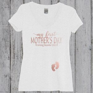 Puede incluir: Camiseta blanca de cuello en V con el texto "my first MOTHER'S DAY - Arriving December 2021" en letras doradas y un par de huellas de beb&eacute; doradas.