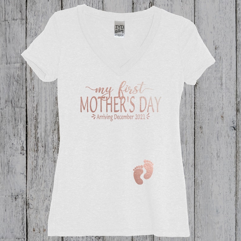 Puede incluir: Camiseta blanca de cuello en V con el texto "my first MOTHER'S DAY - Arriving December 2021" en letras doradas y un par de huellas de beb&eacute; doradas.