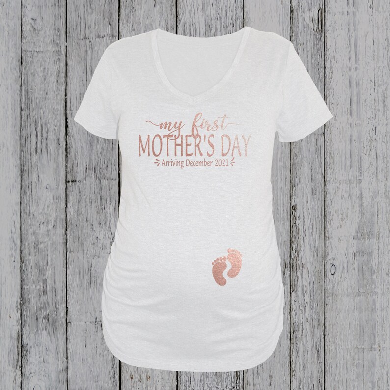 Puede incluir: Una camiseta de maternidad blanca con el texto "my first MOTHER'S DAY" en letras doradas rosas. El texto "Arriving December 2021" tambi&eacute;n est&aacute; en letras doradas rosas. Un par de huellas de pies de beb&eacute; doradas rosas est&aacute;n en la camiseta.