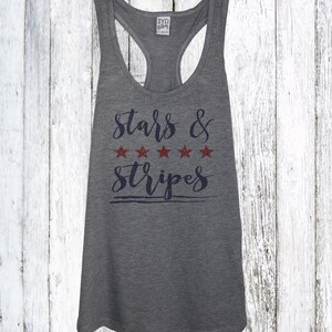 Op de afbeelding: Grijze racerback tanktop met de tekst "Stars & Stripes" in marineblauwe letters en vijf rode sterren. De tanktop heeft een ronde hals en een racerback ontwerp. De achtergrond is een wit houten oppervlak.