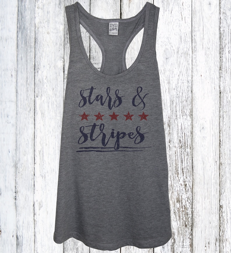 Op de afbeelding: Grijze racerback tanktop met de tekst "Stars & Stripes" in marineblauwe letters en vijf rode sterren. De tanktop heeft een ronde hals en een racerback ontwerp. De achtergrond is een wit houten oppervlak.
