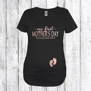 Puede incluir: Camiseta de maternidad negra con el texto "my first MOTHER'S DAY Arriving December 2021" en letras doradas rosas y un par de huellas de pies de beb&eacute; rosas.
