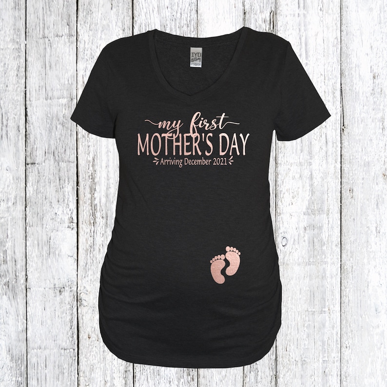 Puede incluir: Camiseta de maternidad negra con el texto "my first MOTHER'S DAY Arriving December 2021" en letras doradas rosas y un par de huellas de pies de beb&eacute; rosas.