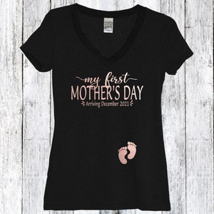 Puede incluir: Camiseta negra de cuello en V con el texto "my first MOTHER'S DAY - Arriving December 2021" en letras doradas y un par de huellas de pies de beb&eacute; doradas.