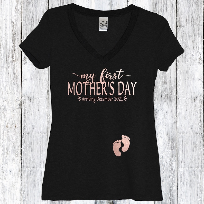 Puede incluir: Camiseta negra de cuello en V con el texto "my first MOTHER'S DAY - Arriving December 2021" en letras doradas y un par de huellas de pies de beb&eacute; doradas.
