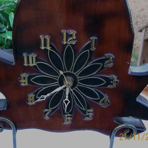 May include: A wooden wall clock shaped like a bird with a black and gold flower design. The clock face has Roman numerals and a black background. The clock is on a stand with a date of 21/11/2012.