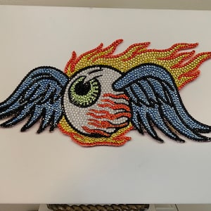 May include: A canvas art piece featuring a stylized eye with blue wings and flames. The eye is white with a green iris and black pupil. The wings are blue, and the flames are red, yellow, and orange. The artwork is embellished with sparkling crystals.