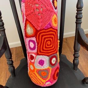May include: A vibrant, handmade crochet pillow in shades of pink, red, orange, and yellow. The pillow features a patchwork design with various geometric shapes and patterns, resting on a dark wooden chair.
